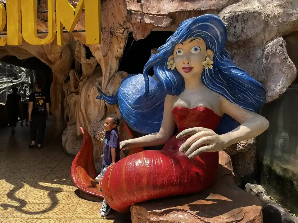 A young boy stands beside a large, colorful statue of a mermaid with blue hair and a red tail, smiling at the camera. The…