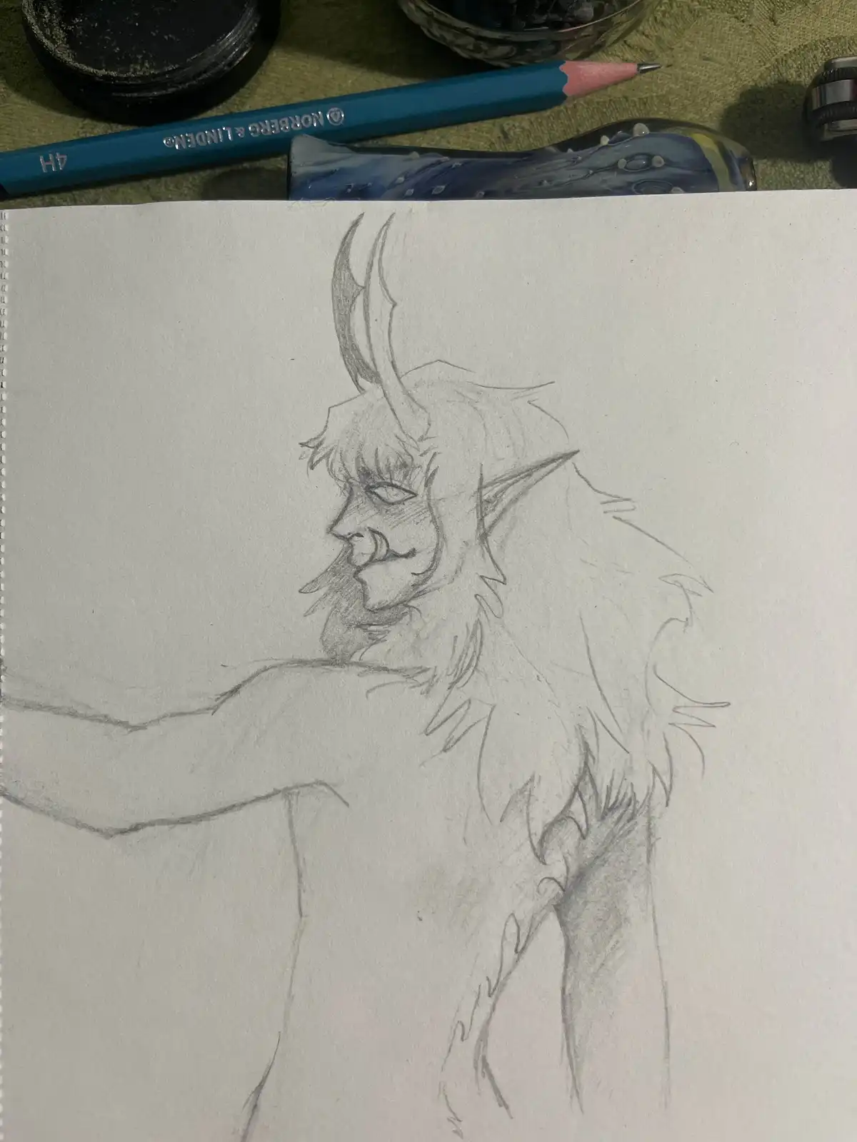 A pencil sketch on a spiral-bound notebook shows a demonic-looking creature with horns, pointed ears, and a beard, drawn in…