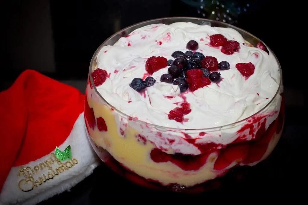 A festive Christmas trifle dessert in a glass bowl, topped with whipped cream and mixed berries, sits beside a red Santa hat…