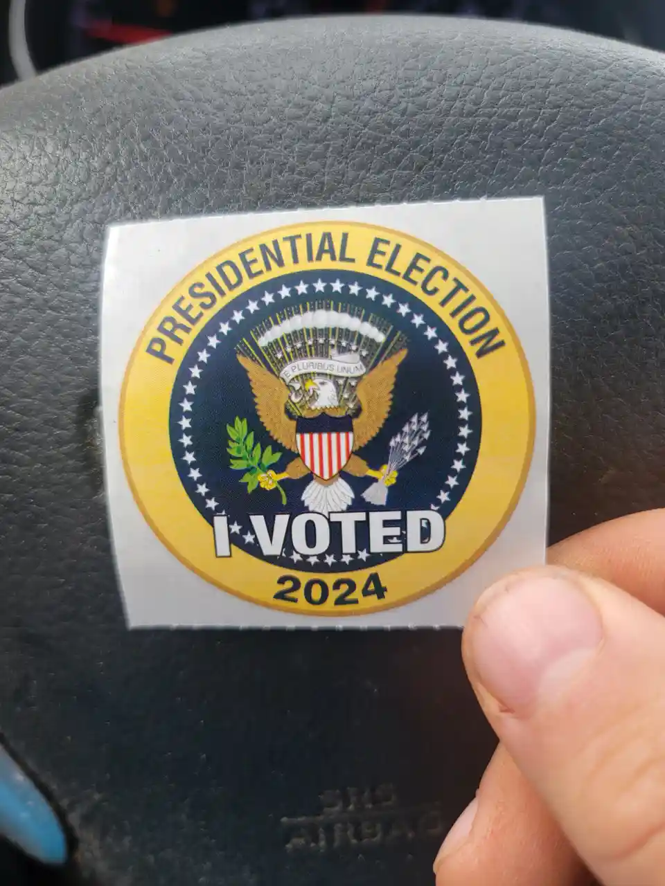A close-up photo shows a hand holding a 2024 U.S. presidential election 'I VOTED' sticker, which features the Great Seal of…