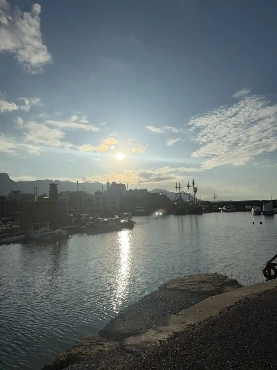 A scenic harbor photo with the sun low in the sky, casting a bright reflection on calm water, surrounded by boats and…