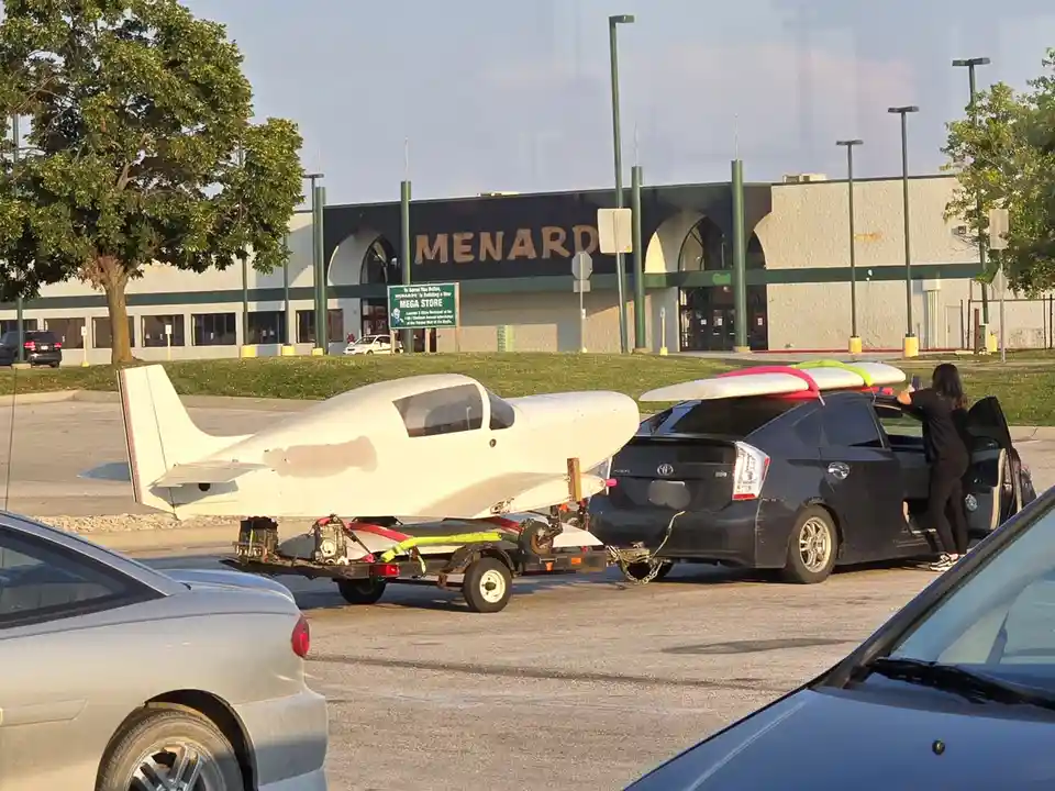 A small white airplane is parked on a trailer in a parking lot in front of a Menards store, next to a black car with…