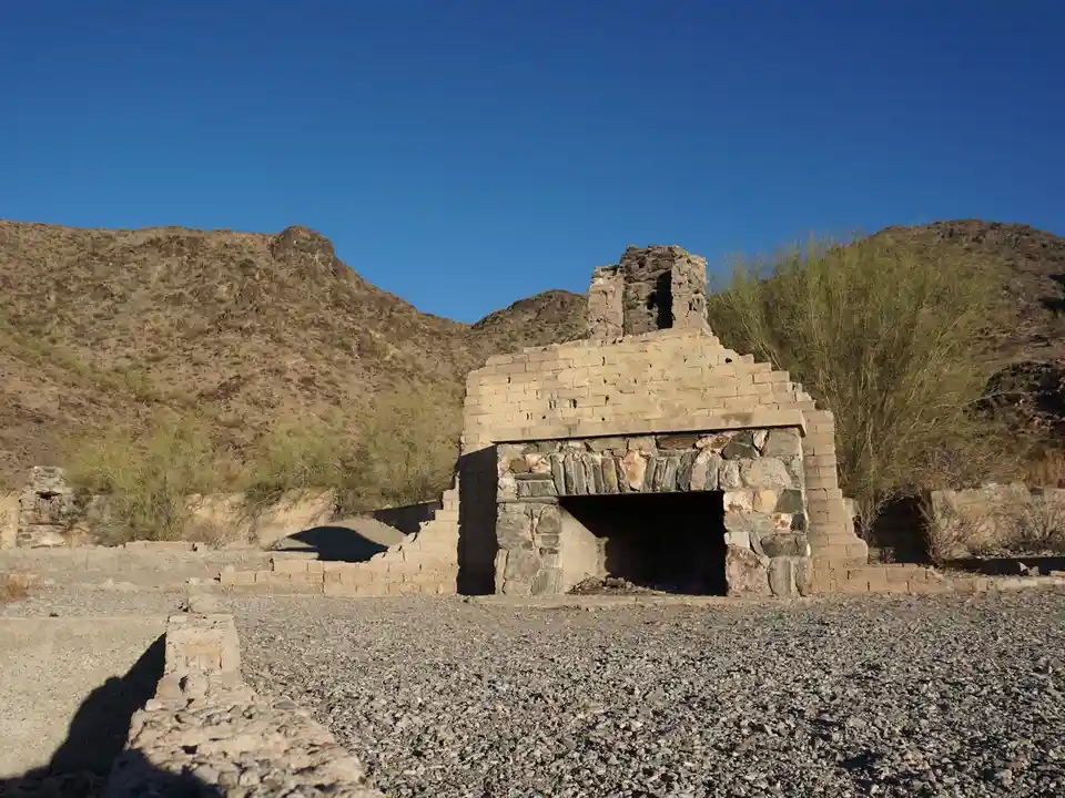 A photograph of a dilapidated stone fireplace ruin in a desert landscape under a clear blue sky, with sparse vegetation and…