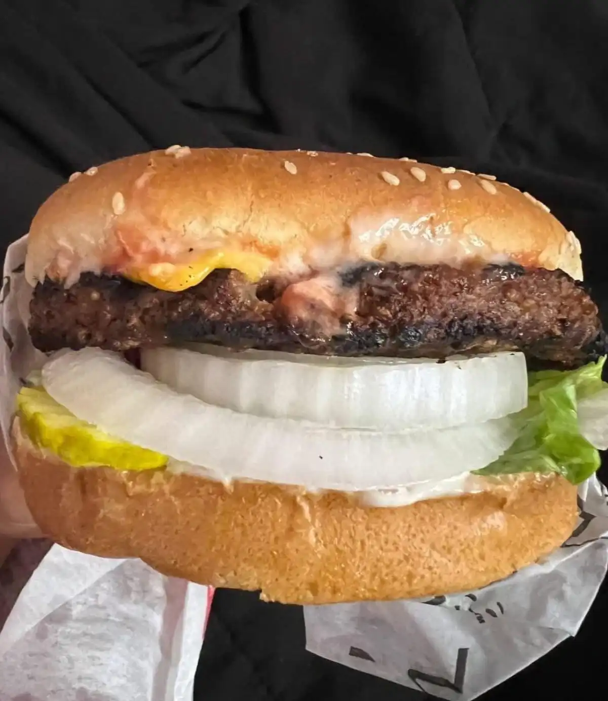 A close-up photo shows a hand holding a cheeseburger with a sesame seed bun, a charred beef patty, melted cheese, multiple…