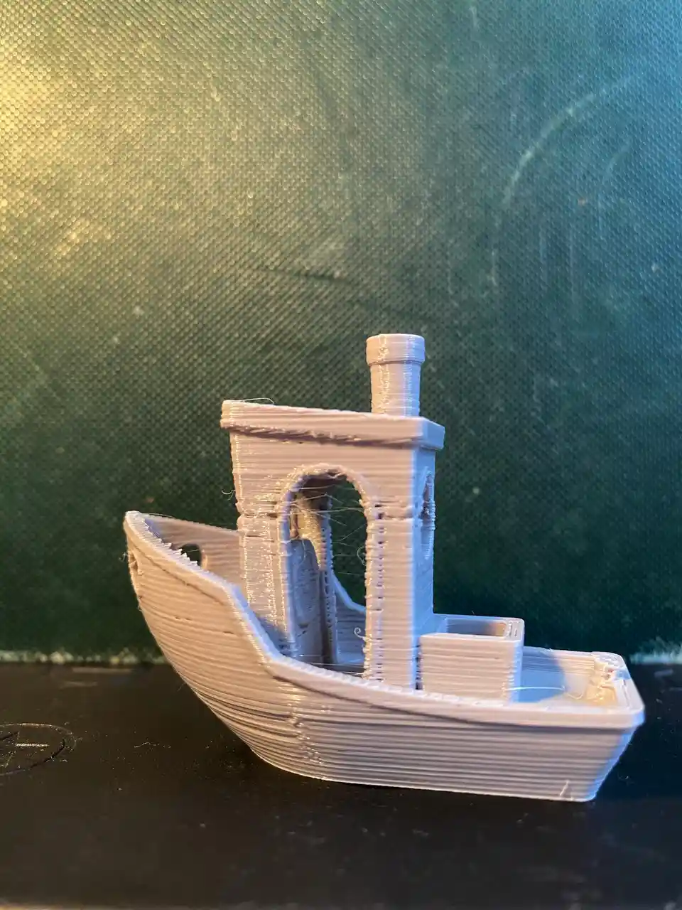 A small, white 3d-printed model boat with visible layer lines sits on a dark surface against a textured green background…