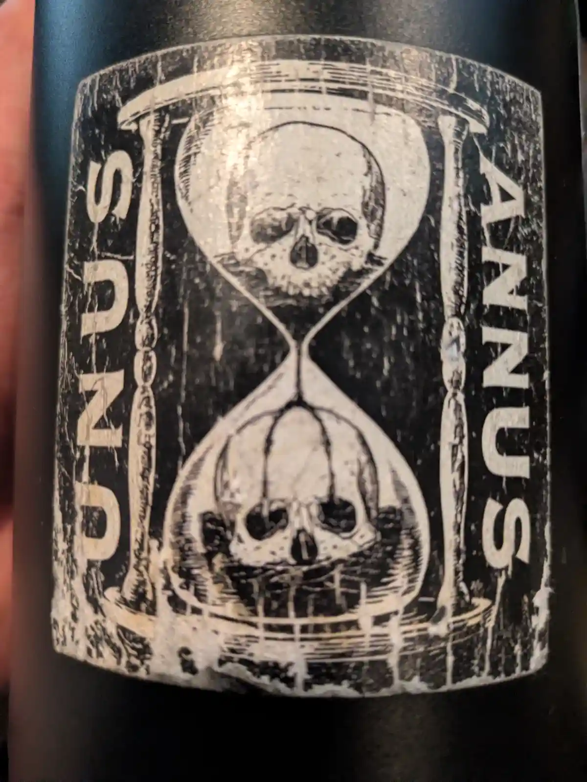 A close-up of a black bottle label featuring a distressed, vintage-style illustration of an hourglass with a skull in each…