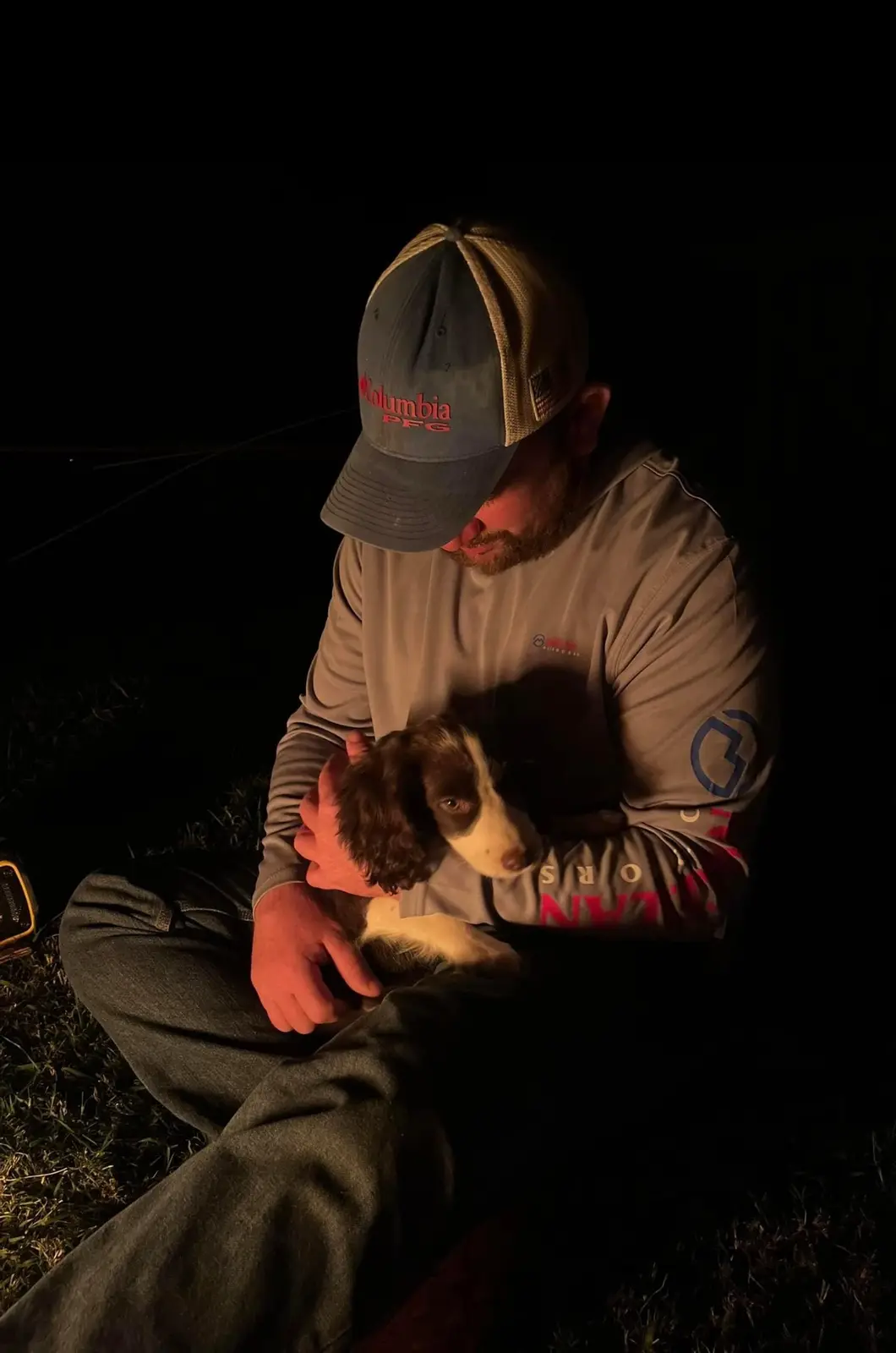 A man wearing a Columbia PFG cap and grey long-sleeve shirt sits in the dark outdoors, holding a small brown and white…