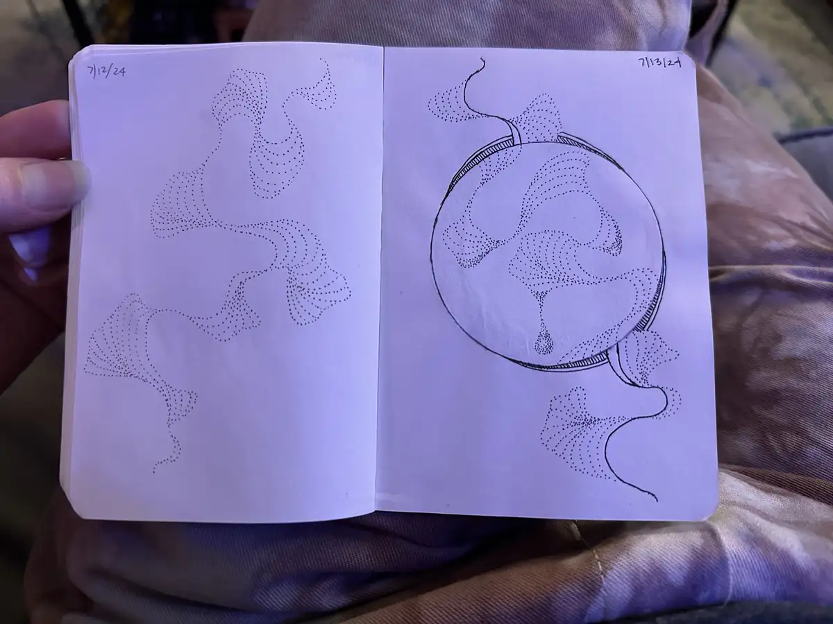 A person holds an open sketchbook showing two pages of black ink dot art drawings. The left page, dated 7/12/24, features…
