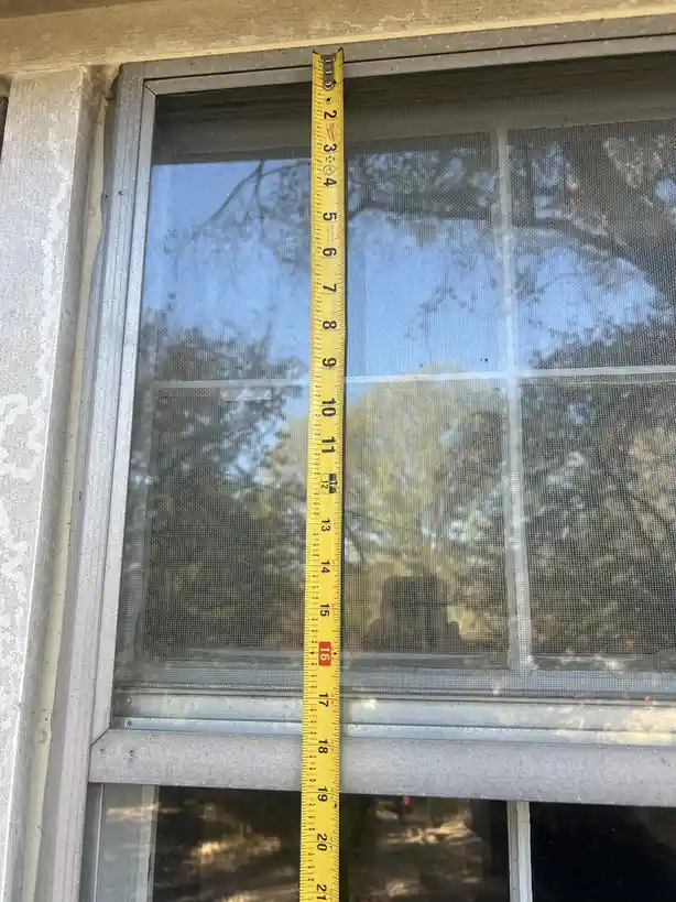 A yellow tape measure is vertically placed against a window screen, showing measurements from 2 to 21 inches. The tape…