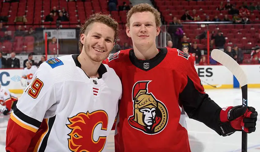Two male ice hockey players pose together on an ice rink, one in a white Calgary Flames jersey with number 9 and the other…