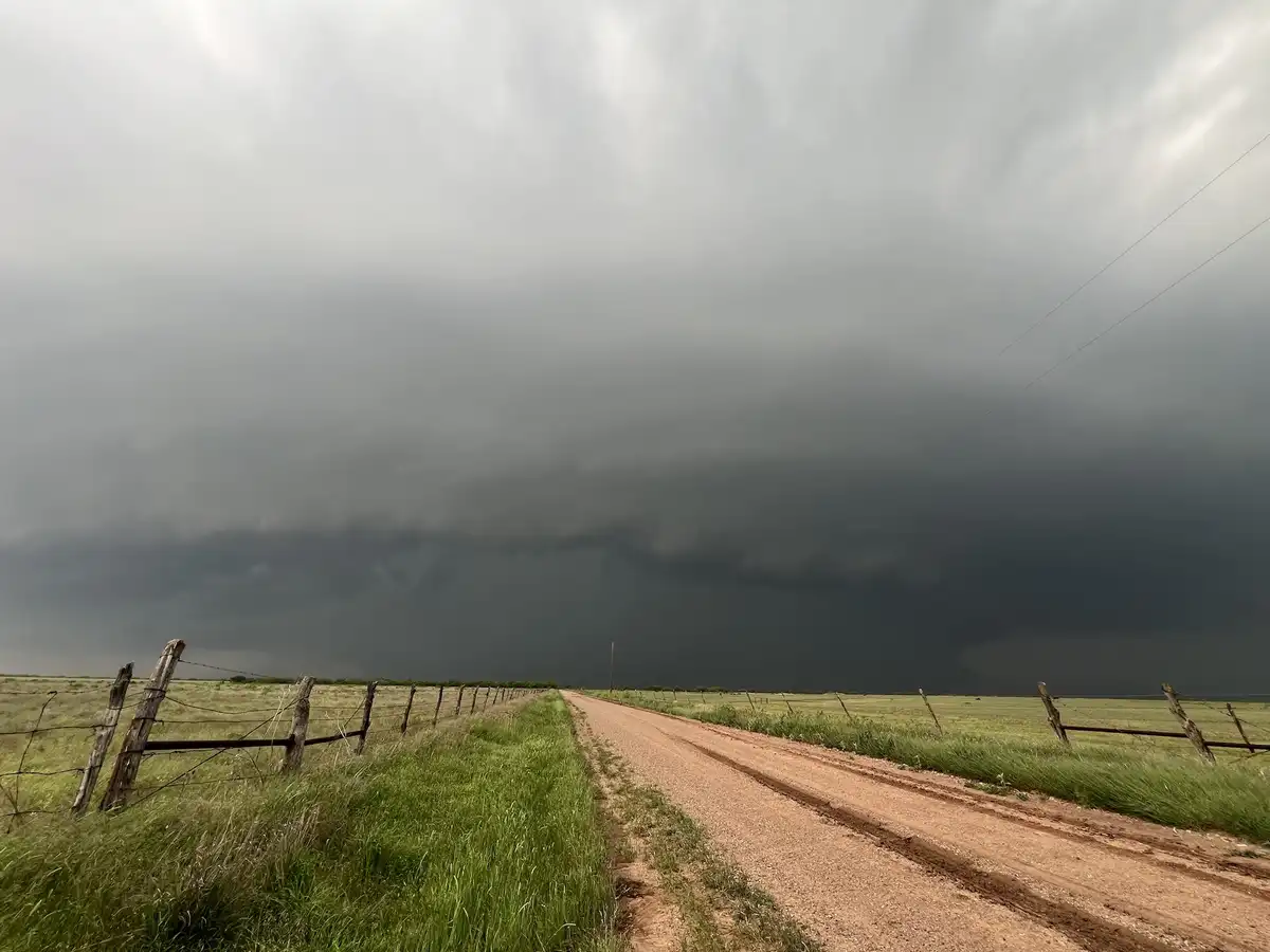 A wide-angle photo shows a dirt road stretching into the distance through a grassy field, flanked by weathered wooden fences…
