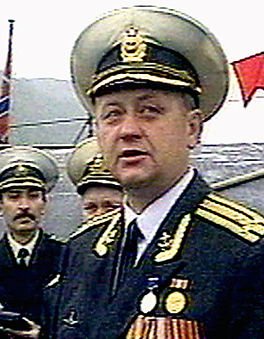 A man in a military uniform with a captain's hat and medals is speaking, with two other men in similar uniforms behind him…