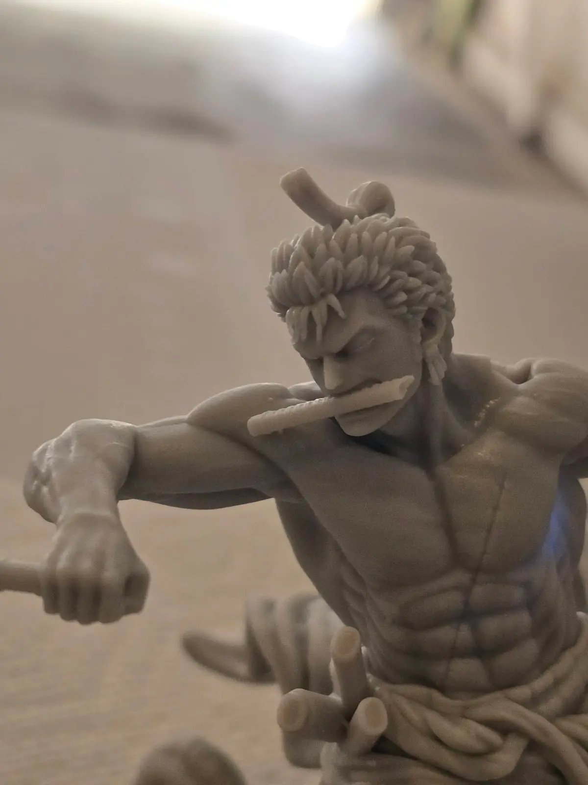 A close-up photo of a grey, un-painted figurine of the anime character Zoro from One Piece, shown in a dynamic fighting pose…
