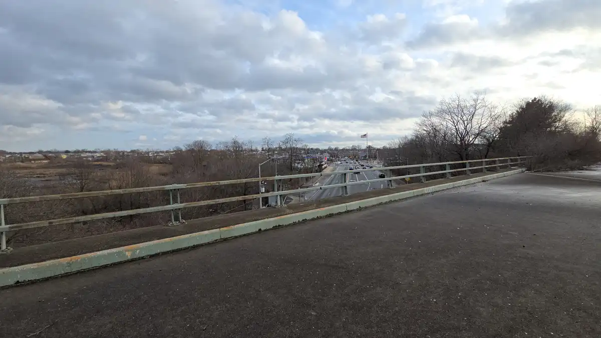 A photo taken from the empty side of a bridge overlooking a busy road with cars, bare trees, and a cloudy sky. The overlaid…