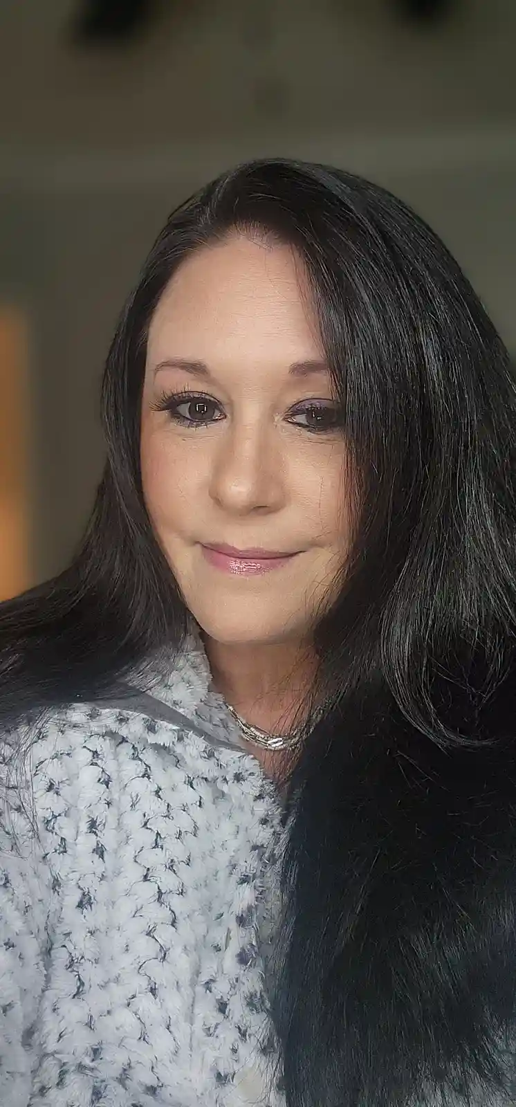 A woman with long dark hair and a subtle smile looks at the camera, wearing a textured gray and black top and a silver…