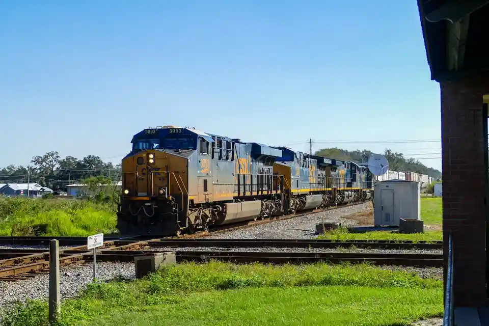 A CSX freight train with locomotive number 3093 travels on tracks under a clear blue sky, passing near a brick building and…