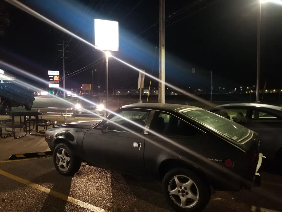 A black classic car, likely a Datsun Z-car, is parked at night in a gas station lot with bright lights causing lens flares…