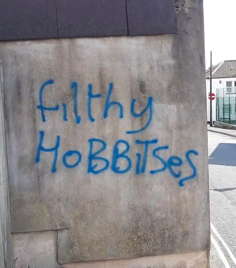 Blue spray-painted graffiti on a concrete wall reads 'filthy HObBITsEs' with inconsistent capitalization. The wall is part…