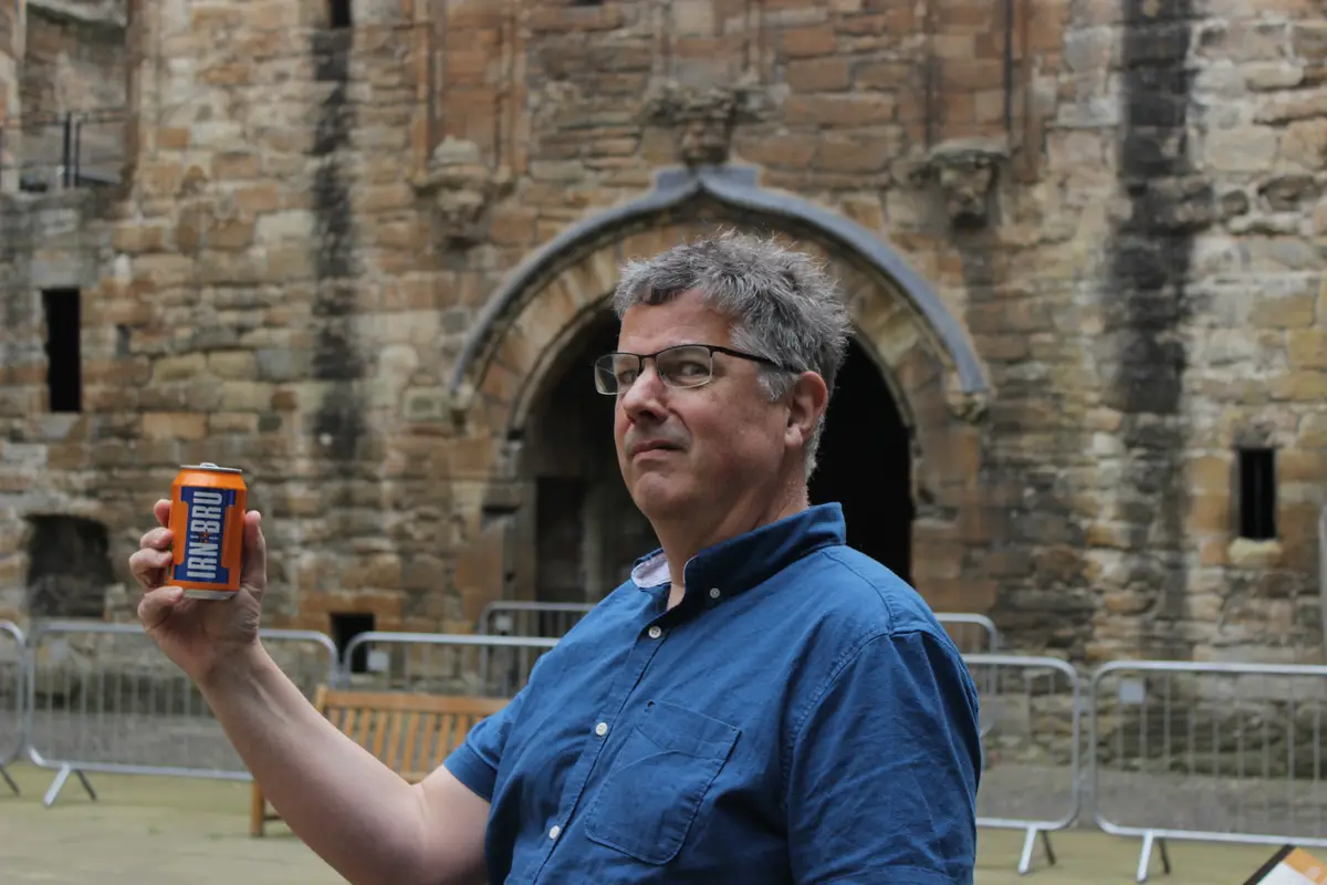 A man in a blue shirt and glasses stands in front of a historic stone building with an arched doorway, holding up an orange…