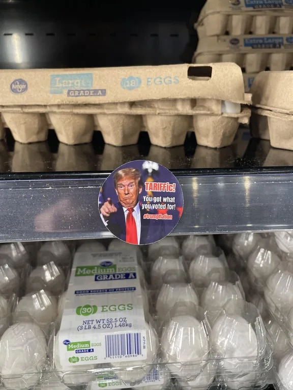 A circular sticker featuring Donald Trump pointing angrily is stuck on a supermarket shelf divider. The sticker has red text…