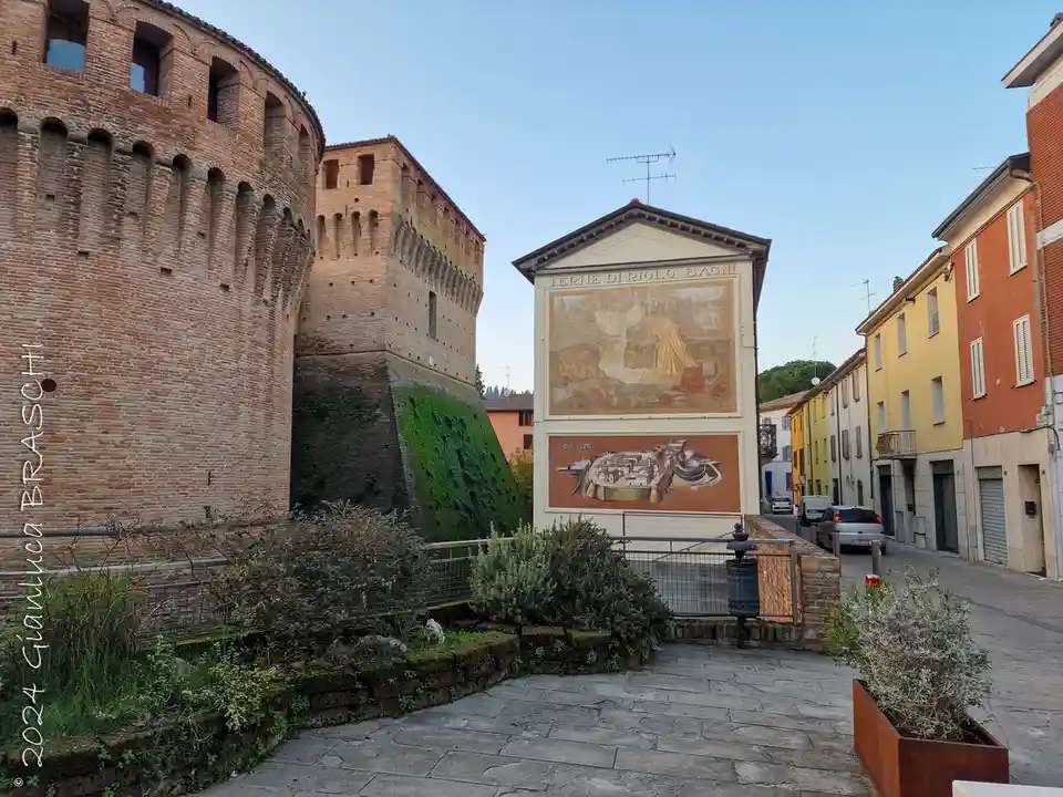 A photo of a historic brick castle with a large mural on a building nearby, showing ancient bath scenes and a city plan. The…