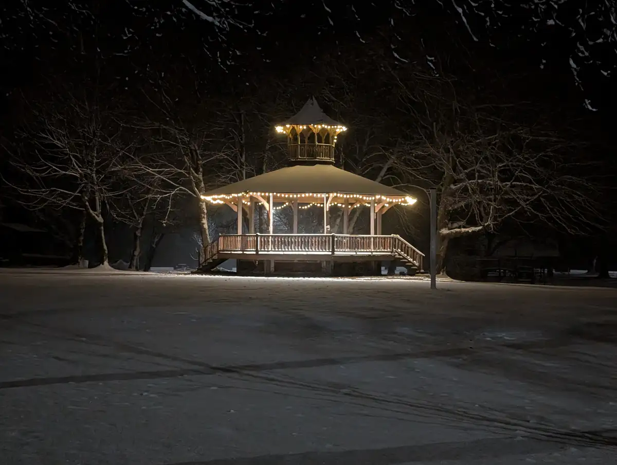 A wooden gazebo in a snowy park at night, illuminated by string lights, surrounded by snow-covered trees and an empty frozen…