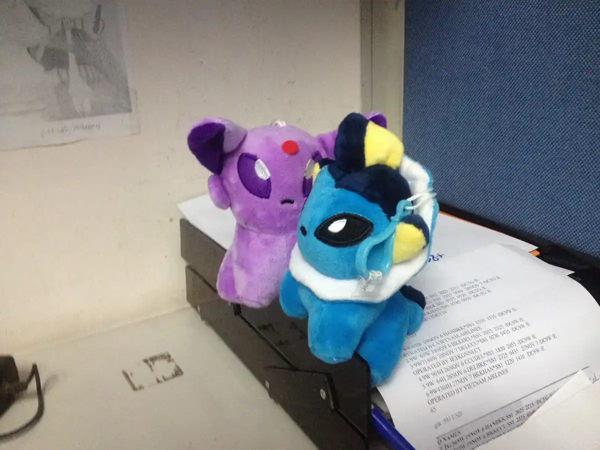 Two plush toys, a purple one and a blue one with a white collar, sit on a desk next to papers. The blue plush has a large…
