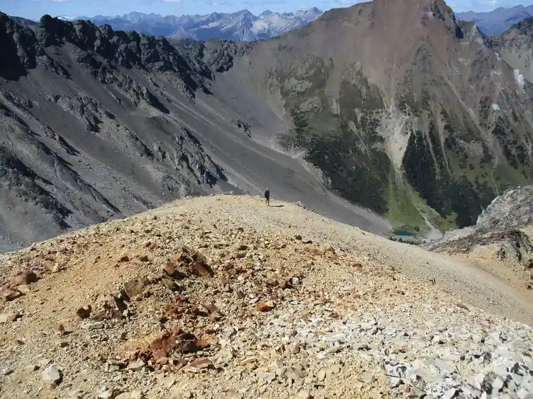 A lone hiker stands on a rocky, barren mountain ridge, dwarfed by vast, rugged peaks and deep valleys under a clear blue…