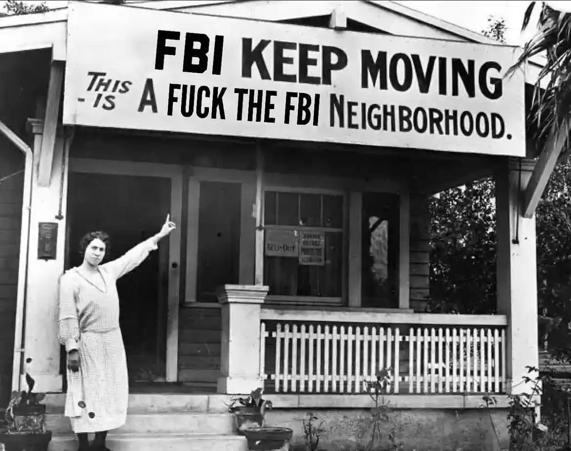 A black-and-white historical photo shows a woman in a dress standing on the steps of a modest house with a white picket…