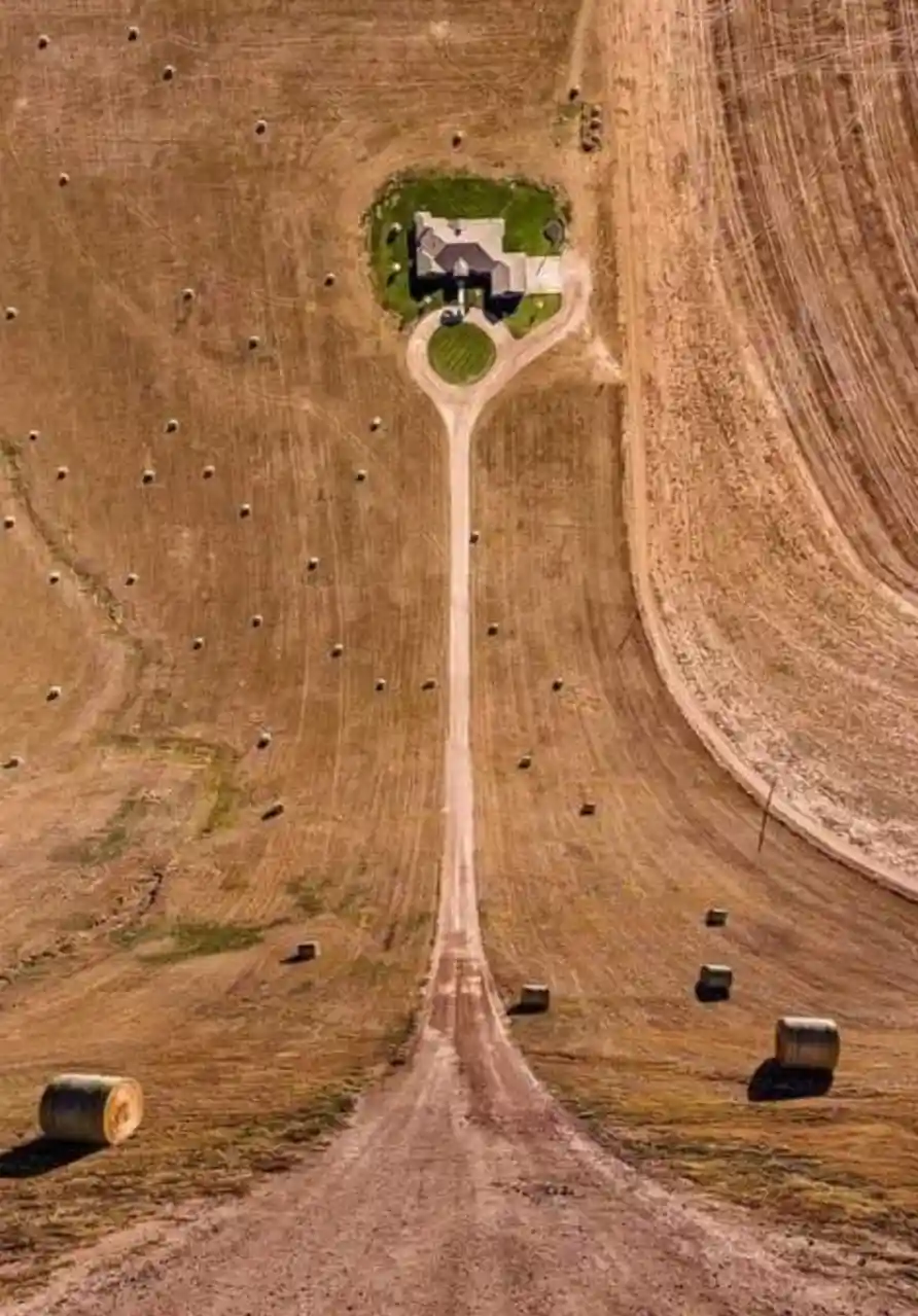 An aerial view shows a single house surrounded by a green lawn and a circular driveway, with a long dirt road leading…