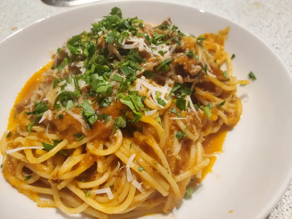 A close-up photo of a white plate filled with spaghetti covered in a rich meat sauce, topped with shredded white cheese and…