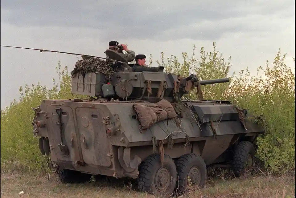 A military armored vehicle with two soldiers on top, one using binoculars and the other looking forward, parked in a grassy…