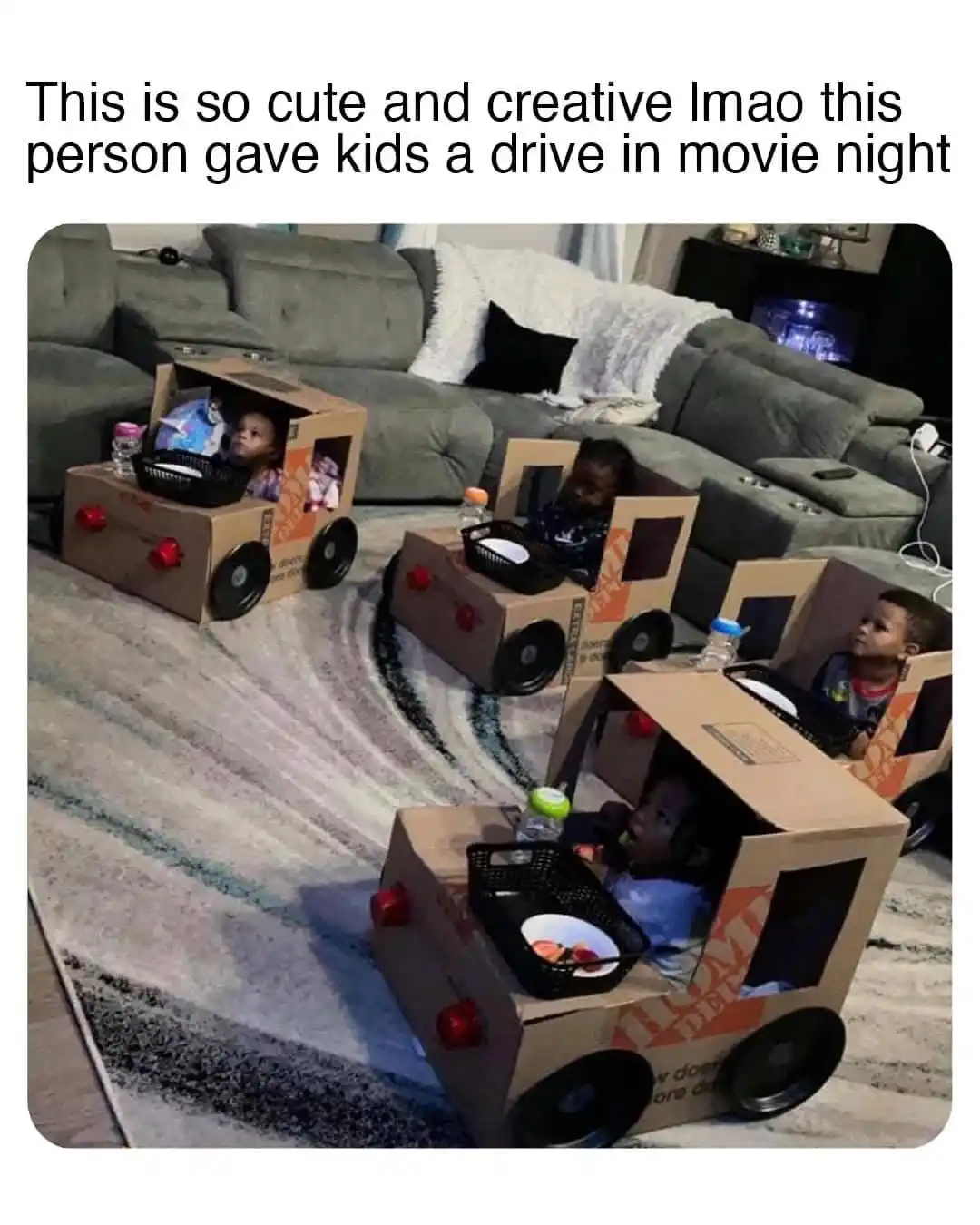 Four children sit inside homemade cardboard cars decorated with wheels and red lights, arranged on a patterned rug in a…