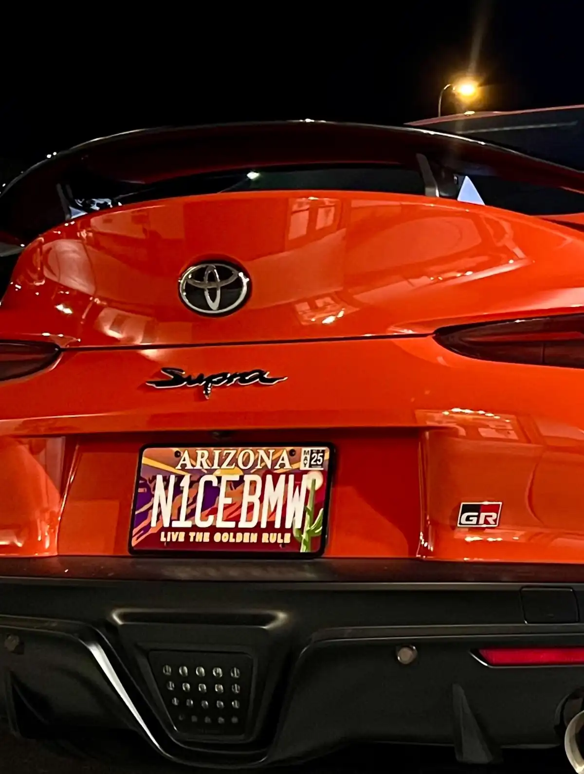 The rear view of a bright orange Toyota Supra at night, featuring a custom Arizona license plate that reads 'N1CEBMW' with…