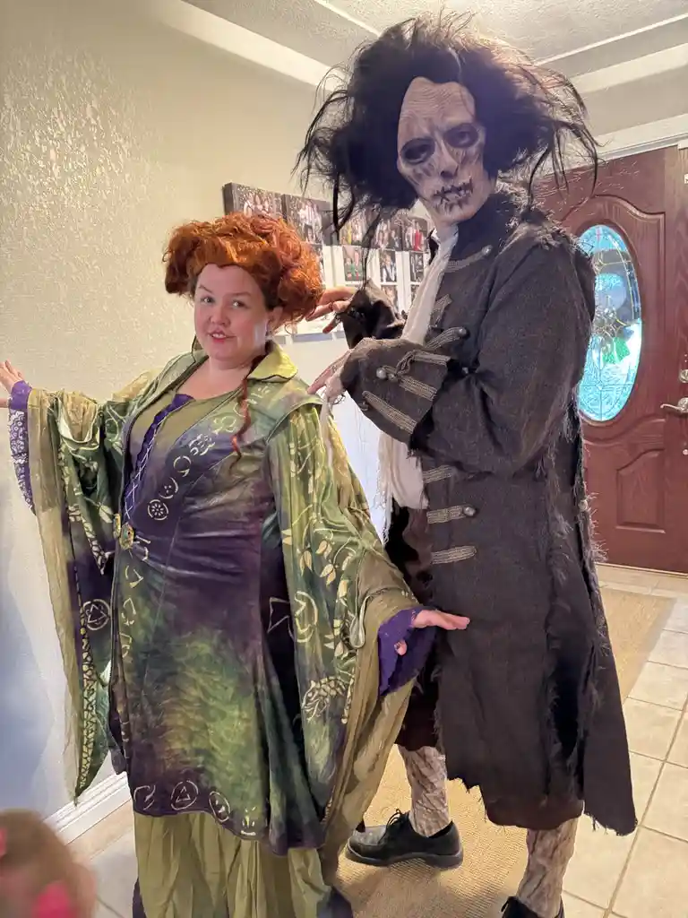 Two people in elaborate costumes stand indoors; one wears a green and purple witch-like robe with a red wig, the other wears…