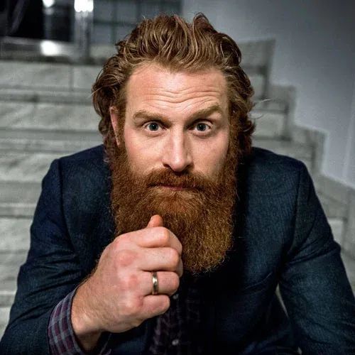 A man with a thick reddish-brown beard and wavy hair, wearing a dark suit and a ring, stares intensely at the camera with…