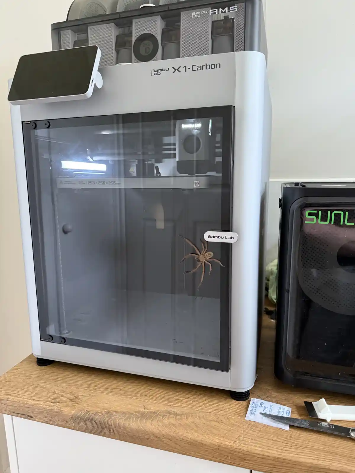 A Bambu Lab X1-Carbon 3D printer with a large brown spider stuck to its glass door, next to a SUNL printer. The overlaid…