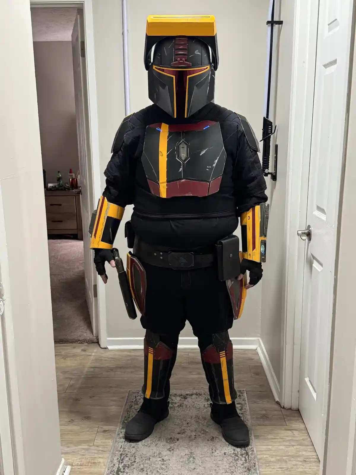 A person in a full-body Mandalorian armor costume, featuring black, yellow, and red coloration, stands in a hallway. The…