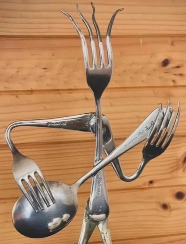 A sculpture made of bent silverware, including forks and a spoon, arranged to resemble a humanoid figure with arms and legs…
