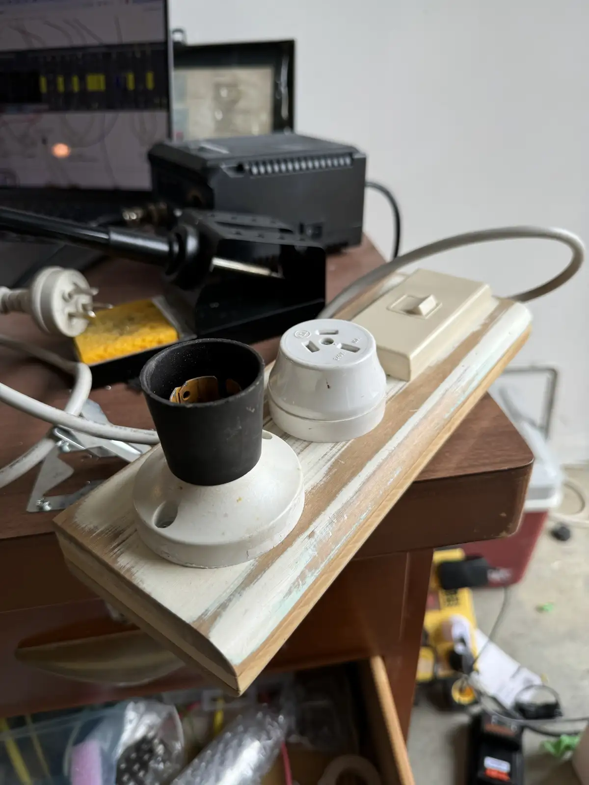 A cluttered workbench holds a white ceramic light socket with its black plastic cap removed, revealing the internal…