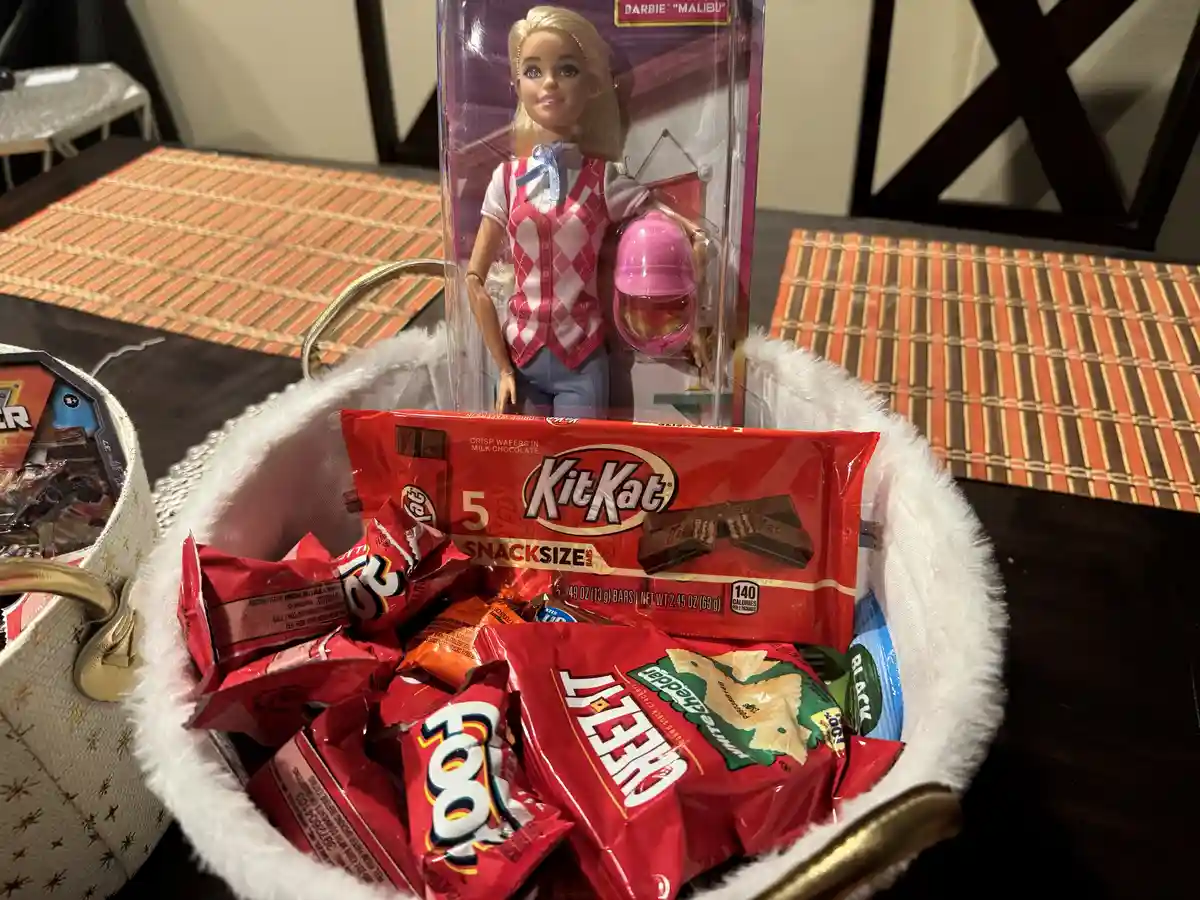 A white, fluffy-lined basket filled with various snack items, including KitKat, Cheetos, and other candies, with a Barbie…
