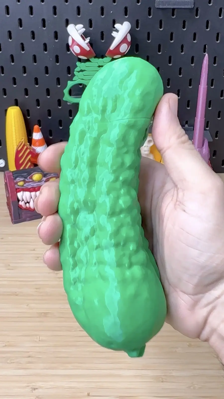 A hand holds a large, green, textured pickle-shaped object against a backdrop of a black pegboard with various colorful toy…