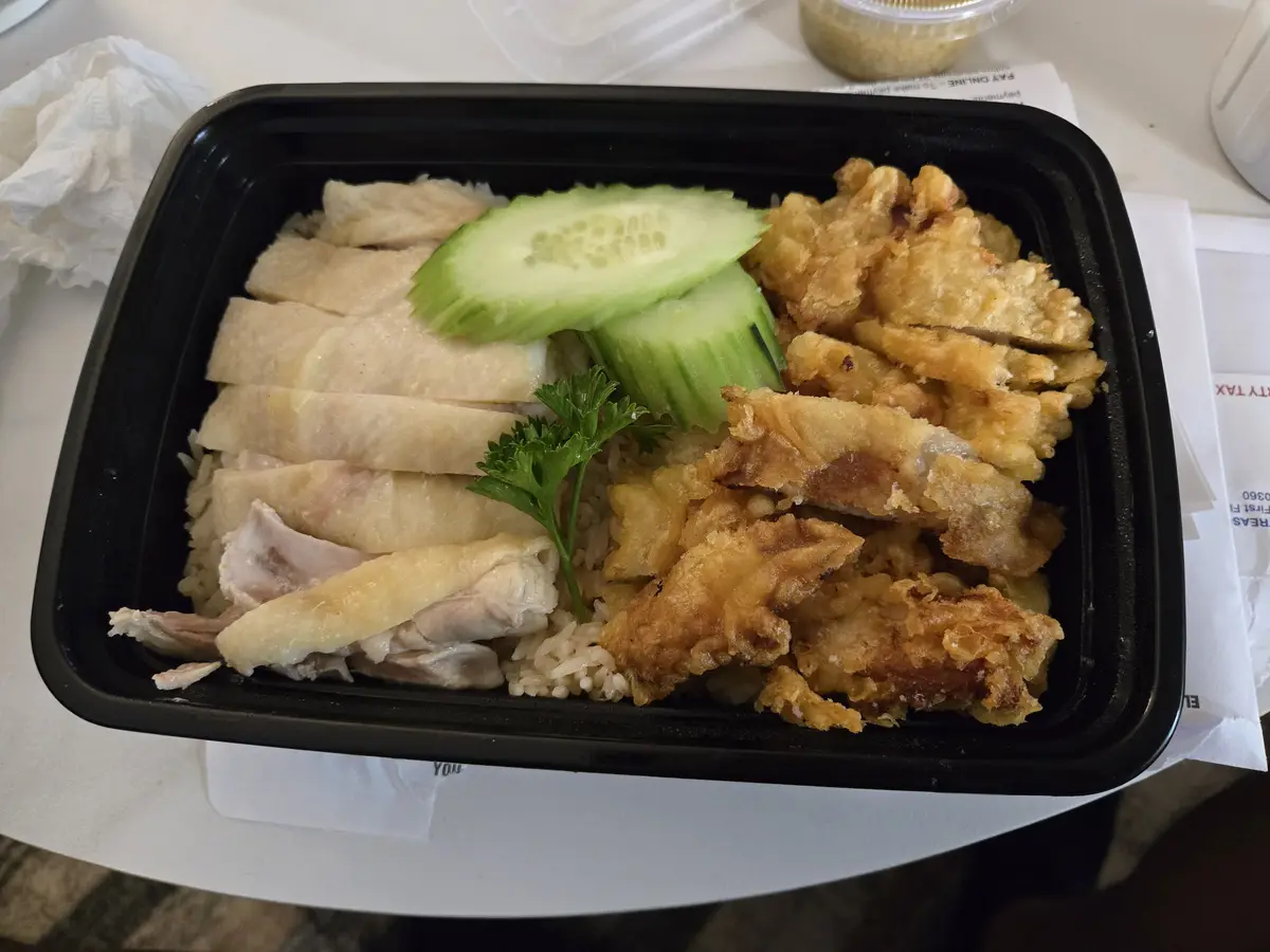 A black takeout container holds a meal with steamed chicken slices, fried chicken pieces, rice, and cucumber slices, with…