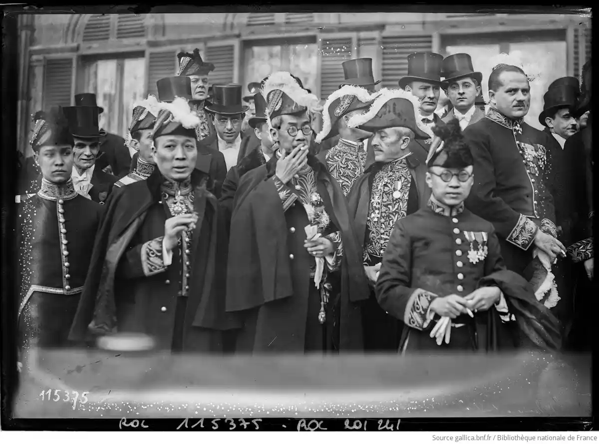 A black-and-white historical photograph shows a group of formally dressed men and women, many wearing ornate hats and…