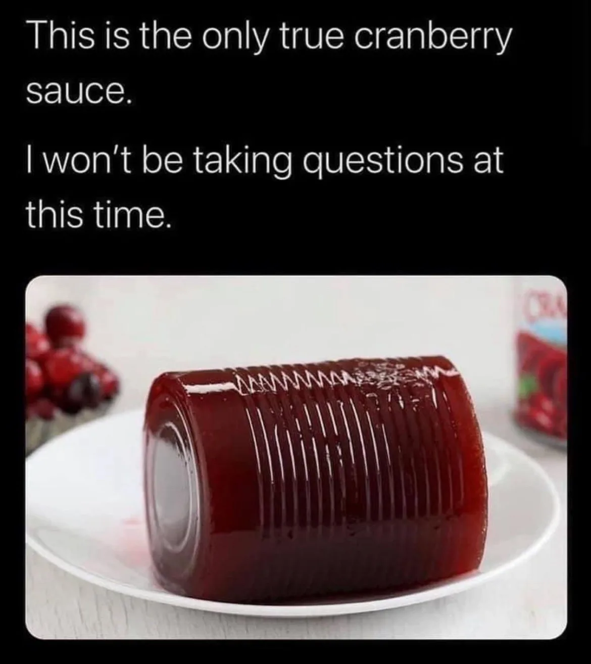 A glossy, deep red, cylindrical cranberry sauce sits on a white plate, its surface showing vertical ridges from being molded…