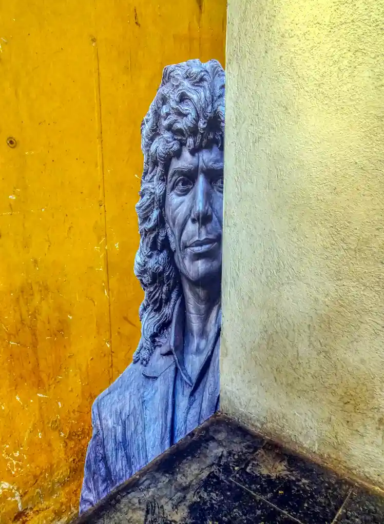 A blue-gray statue of a man with long curly hair and a collared shirt is partially obscured, peeking out from behind a…