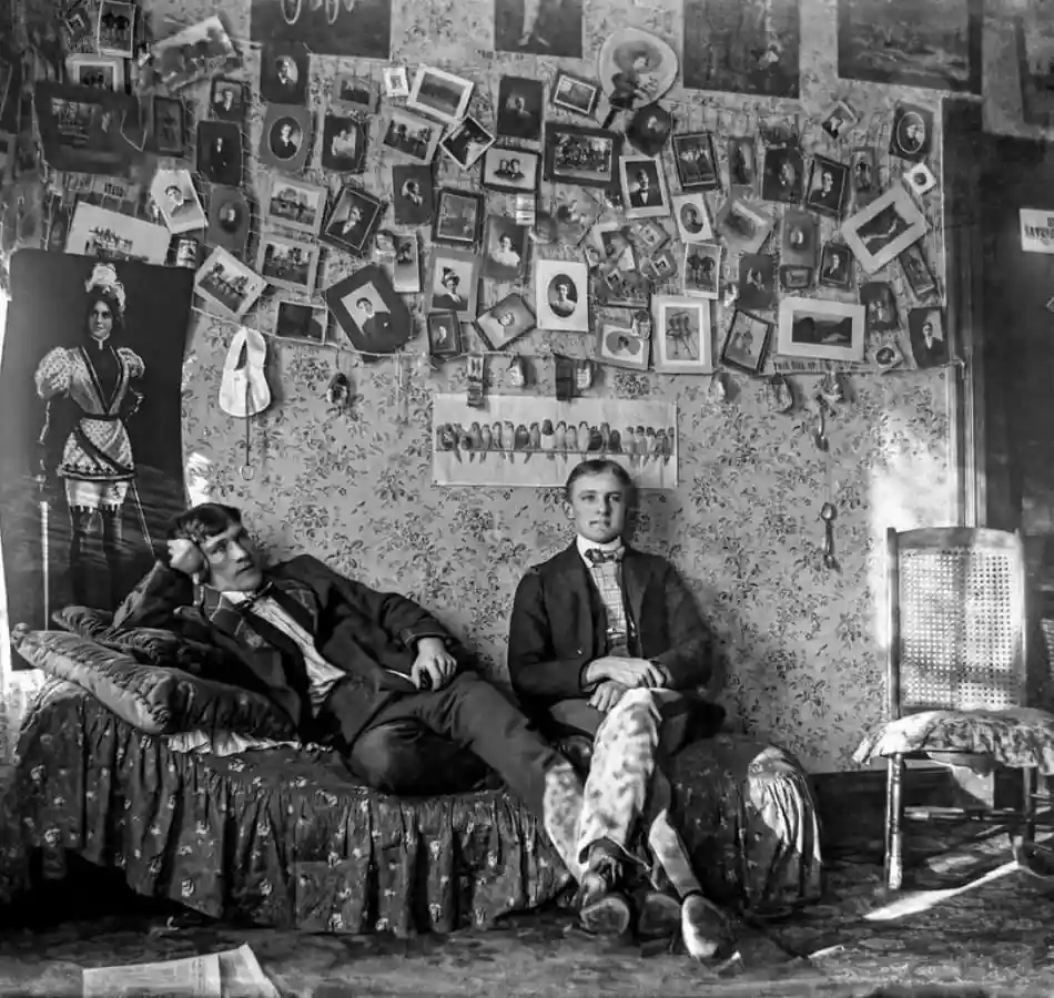 A vintage black-and-white photo shows two young men in a cluttered room; one lounges on a bed looking bored, the other sits…