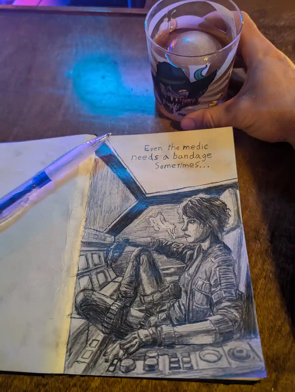 A hand holds a glass with a cartoon demon on it next to a sketchbook on a wooden table. The sketchbook contains a pencil…