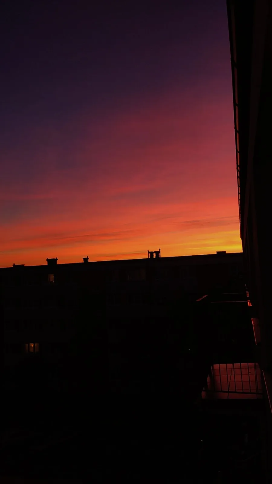 A photograph of a vibrant sunset with deep purple, red, and orange hues over silhouetted apartment buildings. The image is…