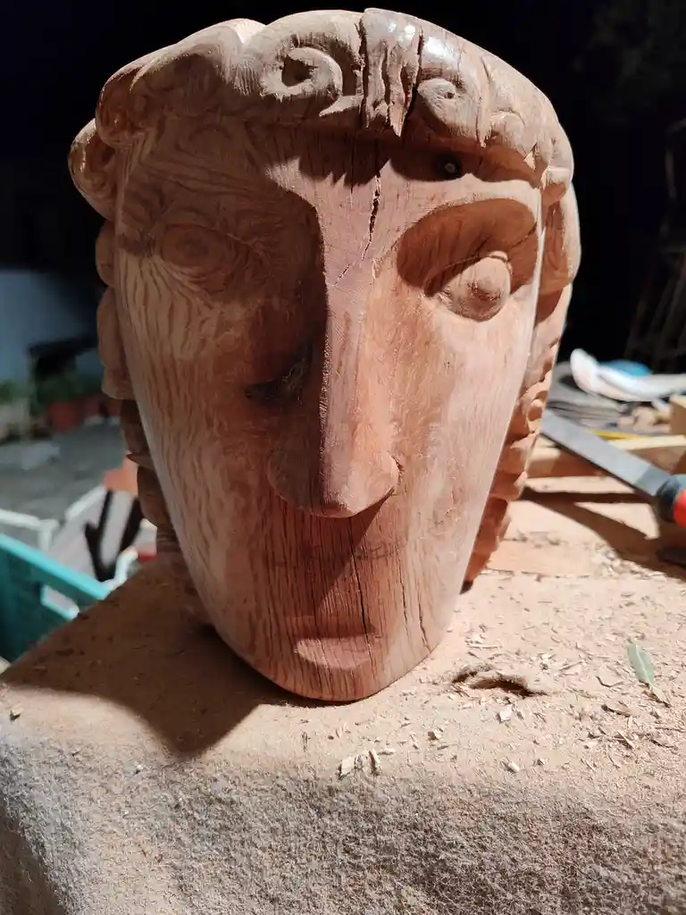 A close-up of a wooden sculpture of a human head, showing visible wood grain and carving marks, resting on a workbench…